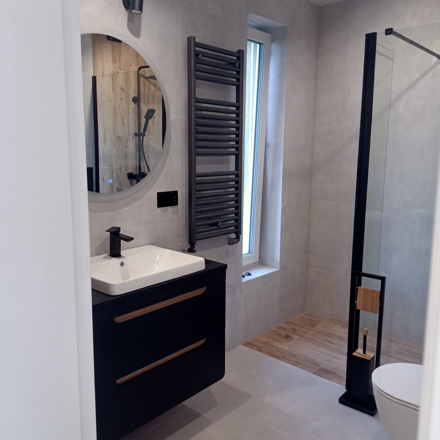 Nowoczesna łazienka z szarymi płytkami, czarnymi akcentami i okrągłym lustrem z podświetleniem LED, widok na kabinę prysznicową ze szklaną ścianką.