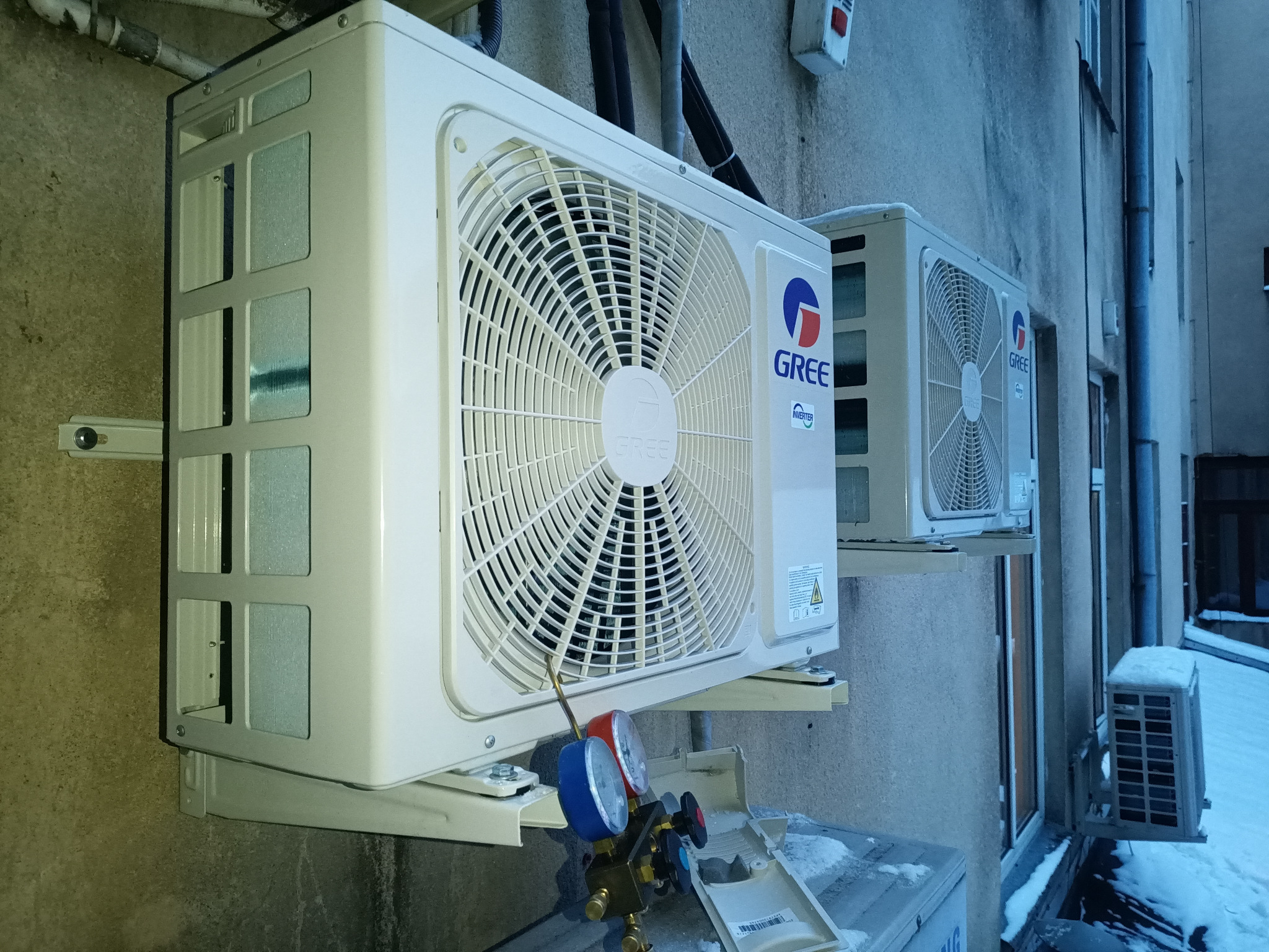 Dwa zewnętrzne moduły klimatyzacji marki Gree zamontowane na ścianie budynku, z widocznymi manometrami serwisowymi podłączonymi do jednego z urządzeń.