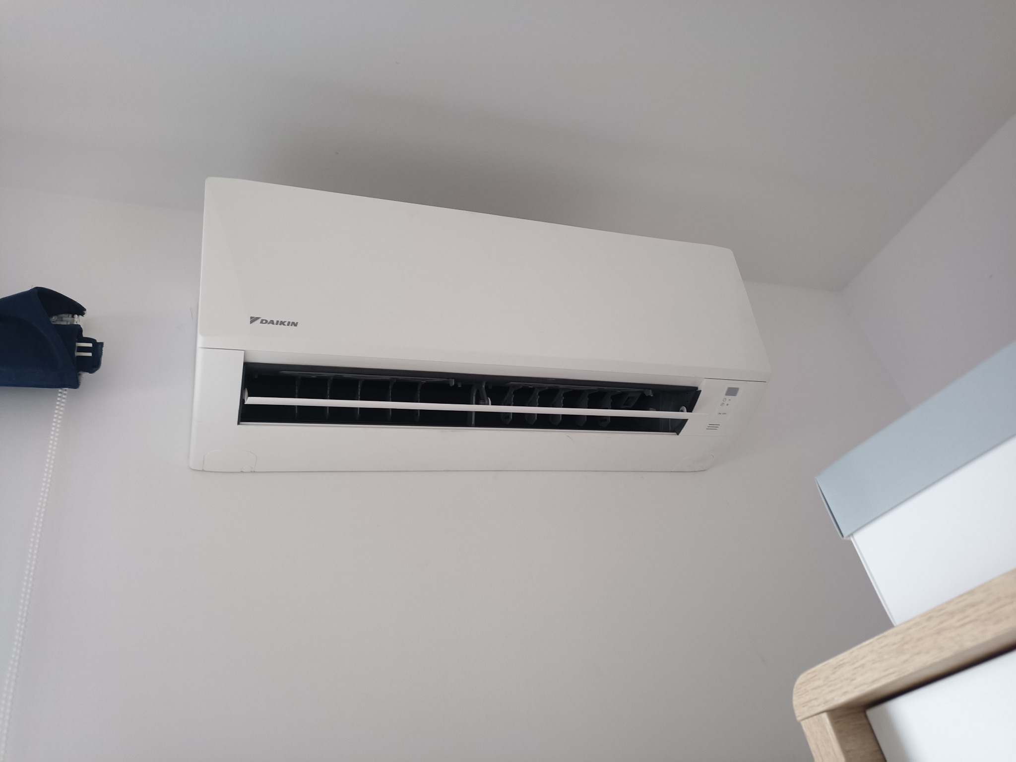 Biała jednostka klimatyzacji Daikin zamontowana na ścianie w jasnym pomieszczeniu, widoczny fragment szafki z szufladami i zasłonięte okno w tle.