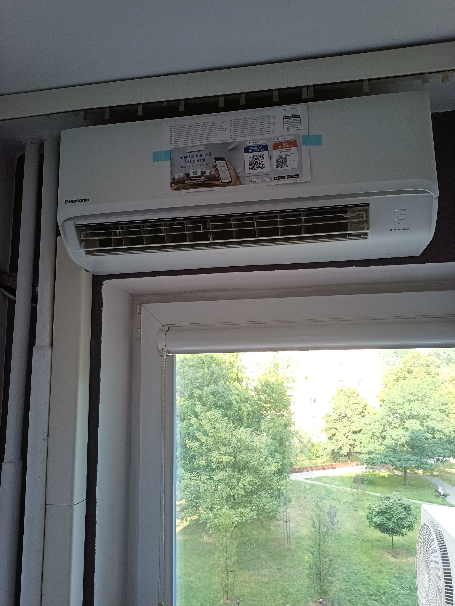 Zamontowana klimatyzacja Panasonic w pomieszczeniu z widokiem na park przez okno. Na urządzeniu naklejka informująca o aplikacji do sterowania.