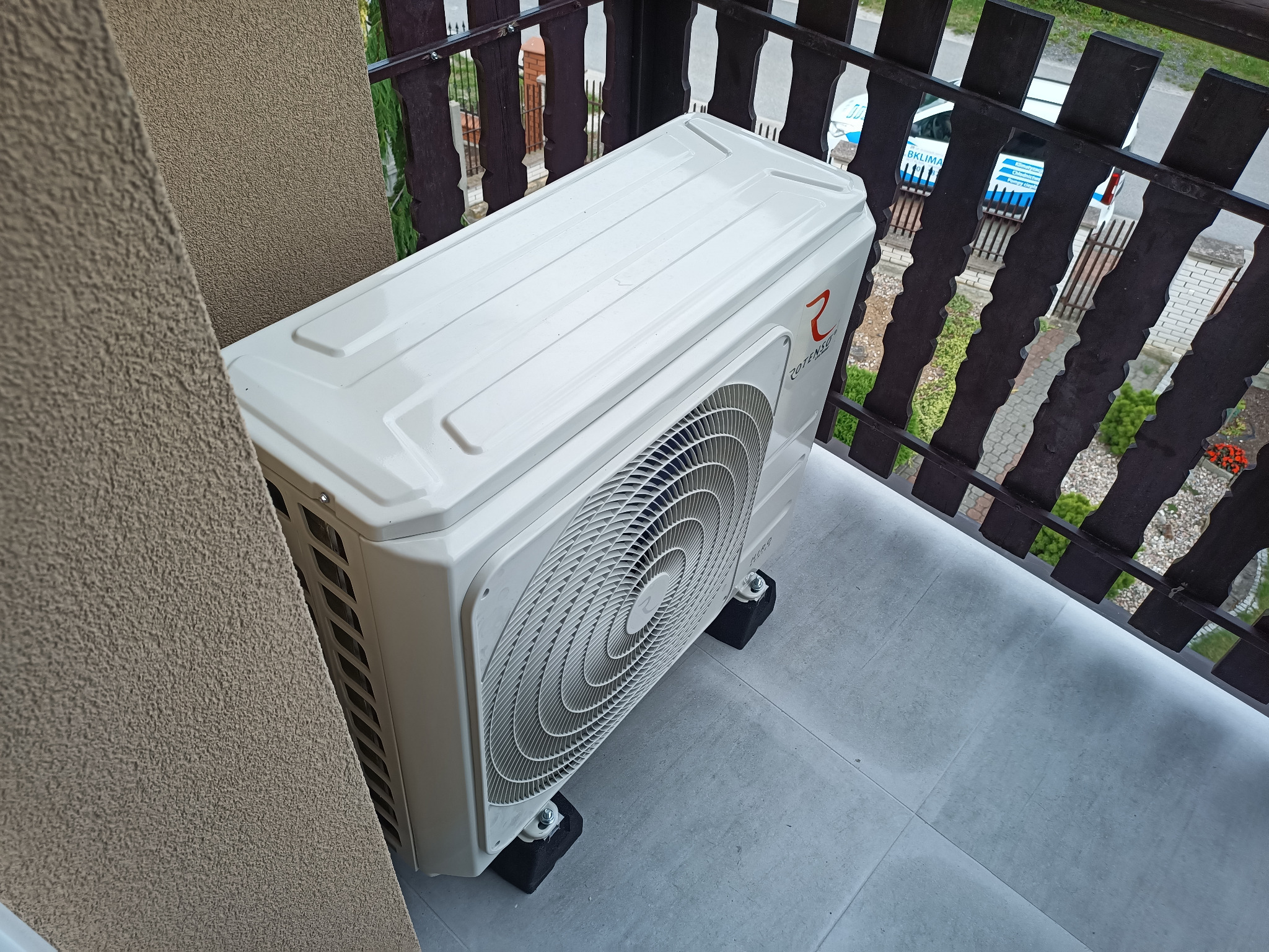 Biała jednostka zewnętrzna klimatyzatora Rotenso umieszczona na balkonie, widoczna z góry, z fragmentem balustrady i elewacji budynku w tle.