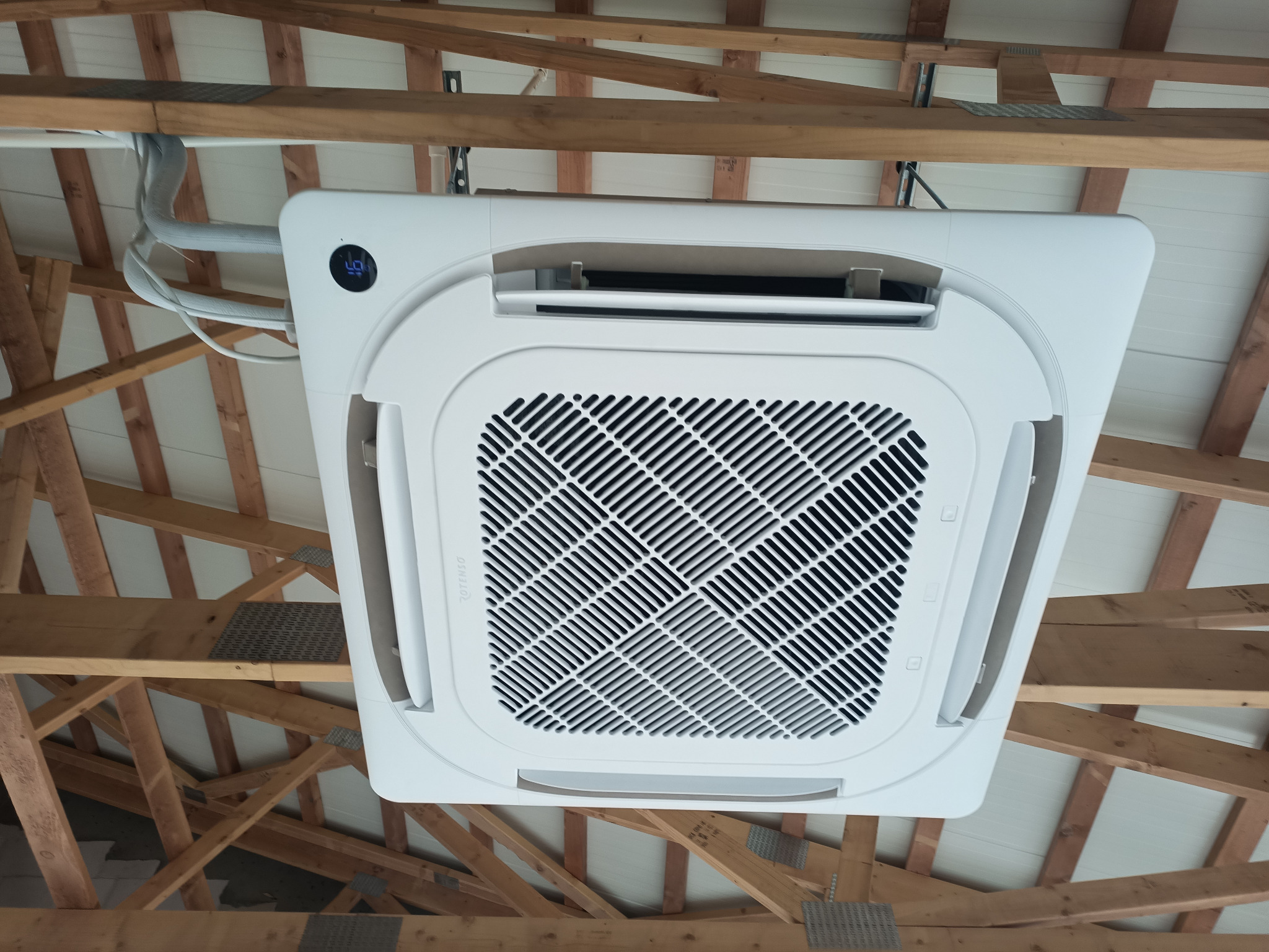 Kasetonowa jednostka klimatyzacyjna zamontowana w suficie podwieszanym z widoczną konstrukcją drewnianą, z wyświetlaczem temperatury w lewym górnym rogu.