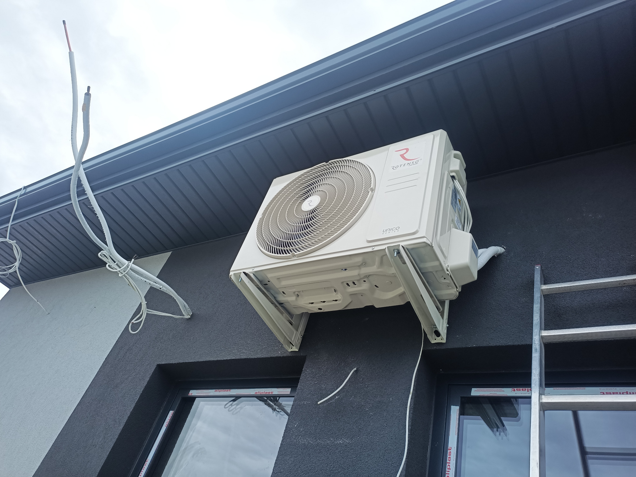 Zewnętrzna jednostka klimatyzacji Rotenso UNICO Series zamontowana na elewacji budynku z widocznymi przewodami i drabina oparta o ścianę.