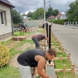 Lakiernia Proszkowa Kewin Radowski - Dwóch mężczyzn w trakcie betonowania fundamentu pod ogrodzenie z czarnymi słupkami. Widoczne drewniane szalunki i narzędzia. Praca w toku, obok droga i dom.