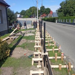 Lakiernia Proszkowa Kewin Radowski - Wykop pod fundament ogrodzenia z drewnianym szalunkiem i czarnymi słupkami, widoczni robotnicy, narzędzia i materiały budowlane. Praca w toku, widok na ulicę i zabudowania.