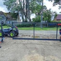 Lakiernia Proszkowa Kewin Radowski - Montaż przesuwnej bramy wjazdowej z siatki na posesji. Obok quad i narzędzia. Prace instalacyjne w toku, widoczna sylwetka montera. Tło: domy i zieleń.