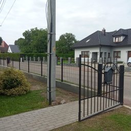 Lakiernia Proszkowa Kewin Radowski - Ukończony montaż ogrodzenia panelowego z bramką i skrzynką na listy. W tle widoczny dom jednorodzinny i dwóch mężczyzn przy pracy.