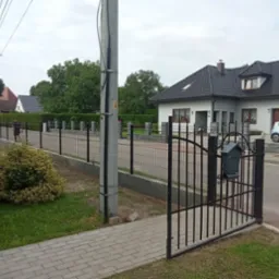 Ukończony montaż ogrodzenia panelowego z bramką i skrzynką na listy. W tle widoczny dom jednorodzinny i dwóch mężczyzn przy pracy.