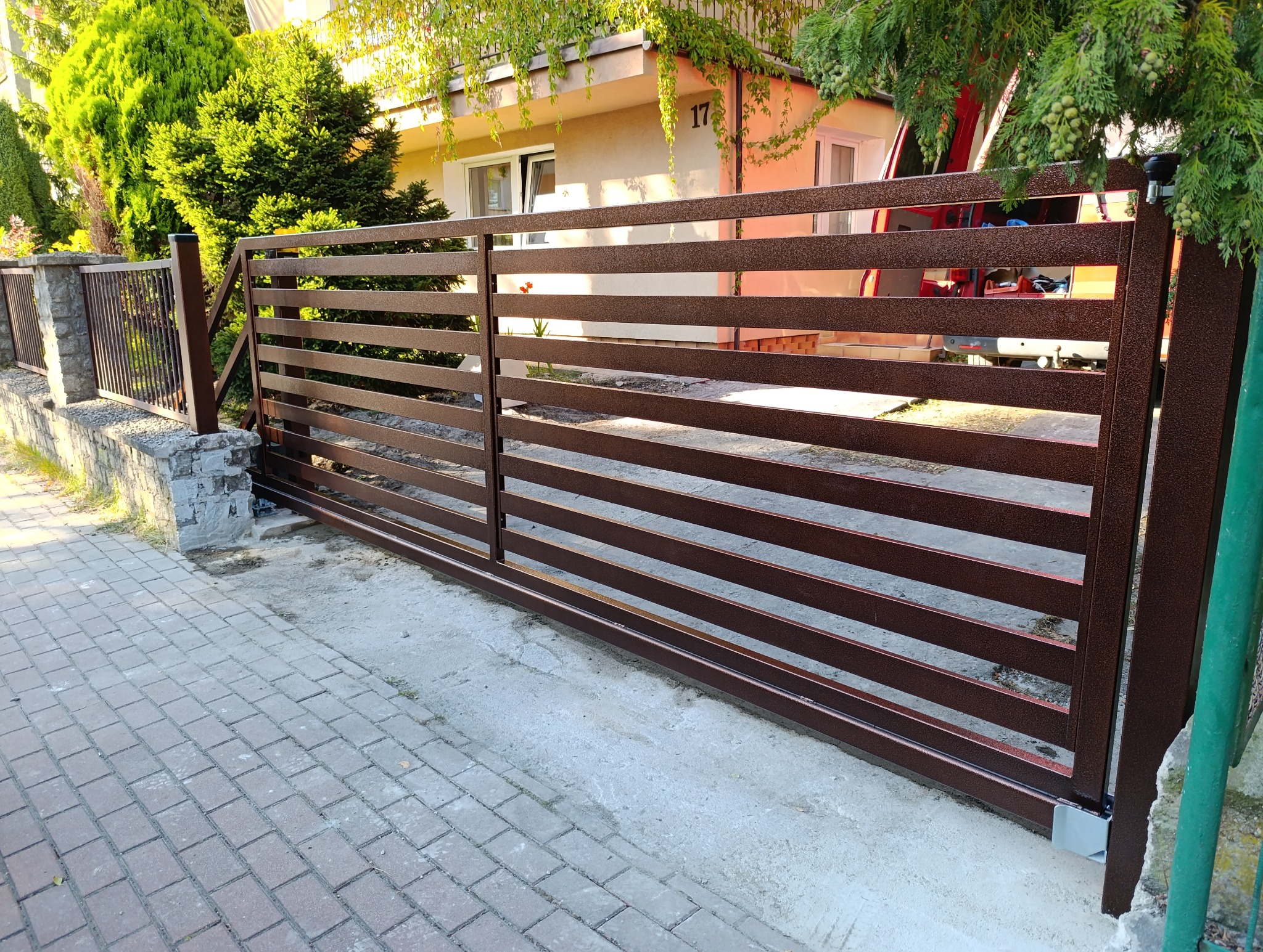 Nowoczesna brama wjazdowa przesuwna z poziomych, metalowych profili w kolorze brązowym, zamontowana przed domem z numerem 17, widok z zewnątrz.