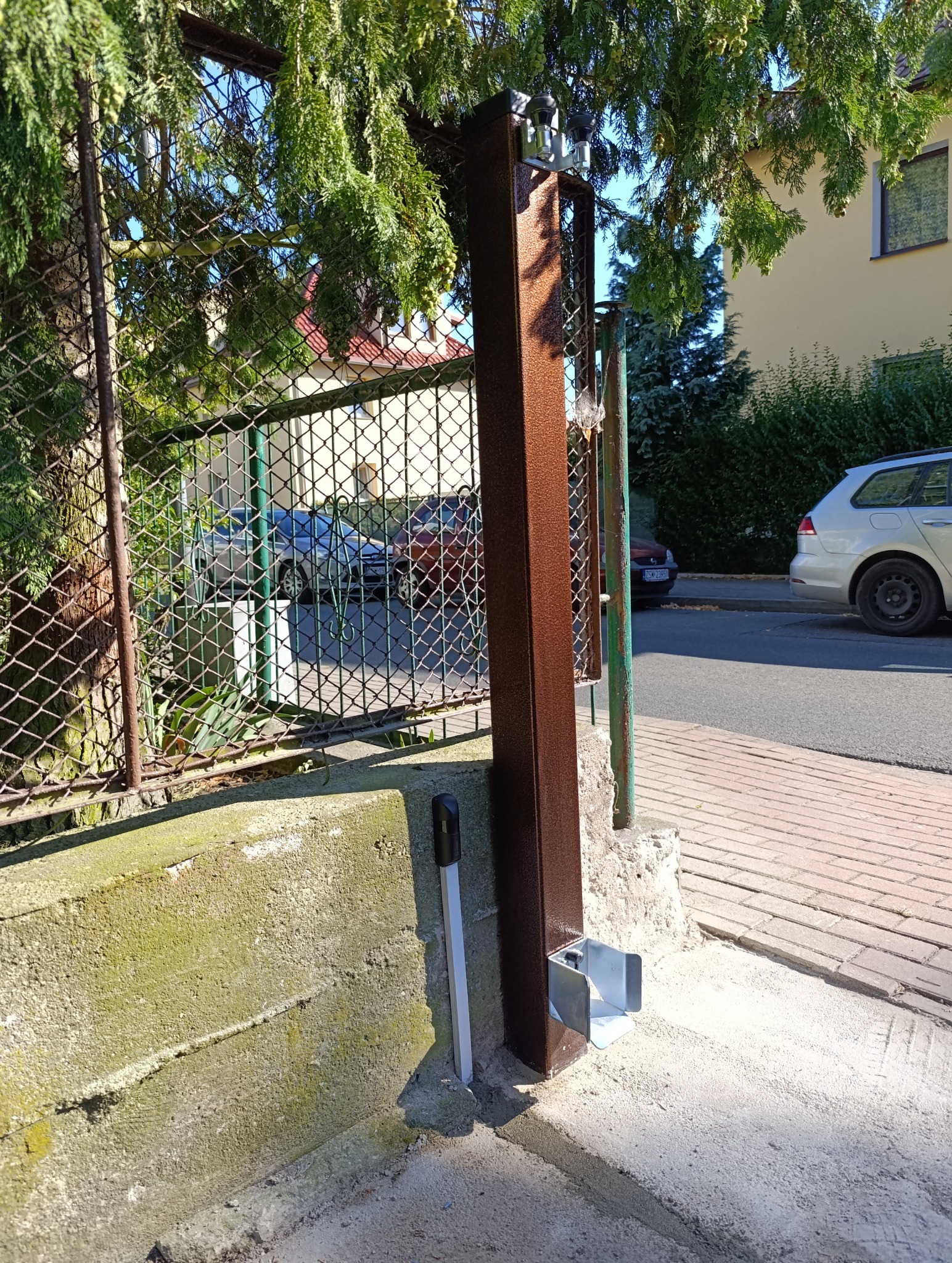 Fragment montażu słupka bramy wjazdowej z automatyką, widoczny na tle istniejącego ogrodzenia z siatki i zieleni.
