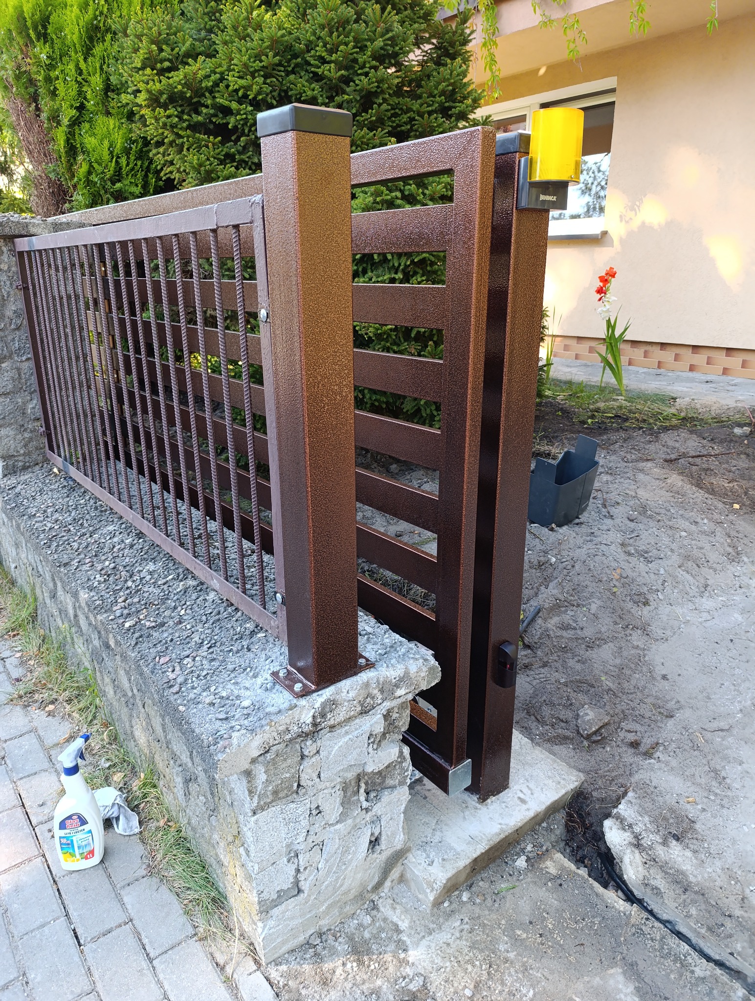 Częściowo zamontowana brama wjazdowa w kolorze miedzianego brązu, z widocznym napędem i elementami ogrodzenia, na tle zieleni.