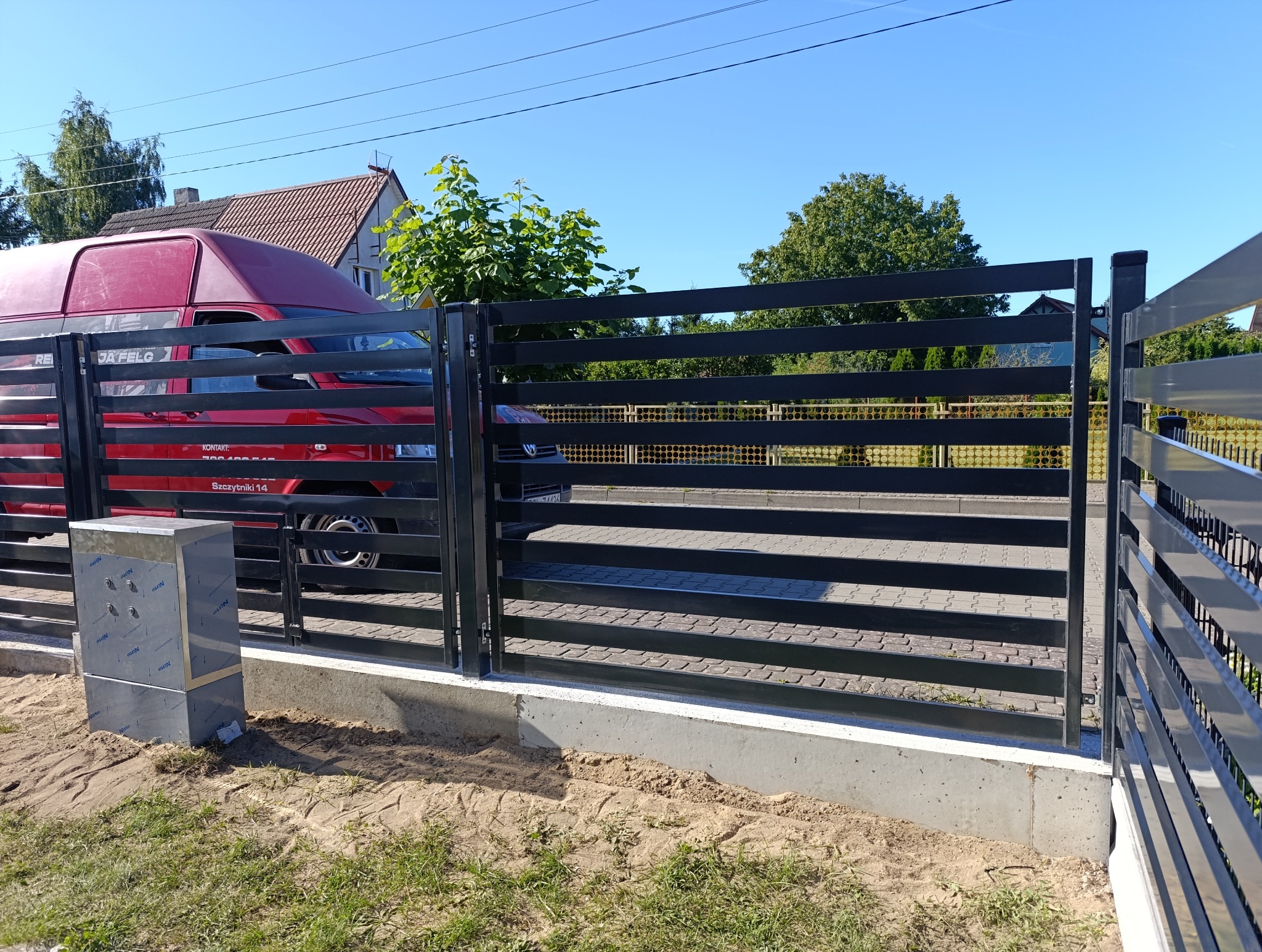 Nowoczesne ogrodzenie metalowe z poziomymi przęsłami w kolorze antracytowym, na betonowej podmurówce. W tle czerwony samochód dostawczy i zieleń. Słoneczny dzień.