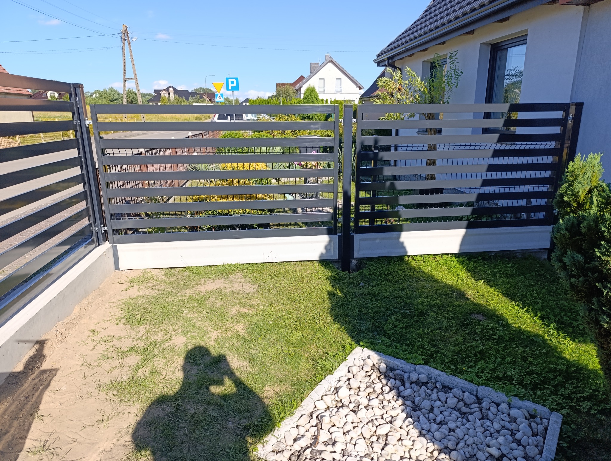 Nowoczesna brama wjazdowa z poziomych, szarych profili, osadzona na betonowej podmurówce. Za bramą widoczny ogród i zabudowa jednorodzinna.