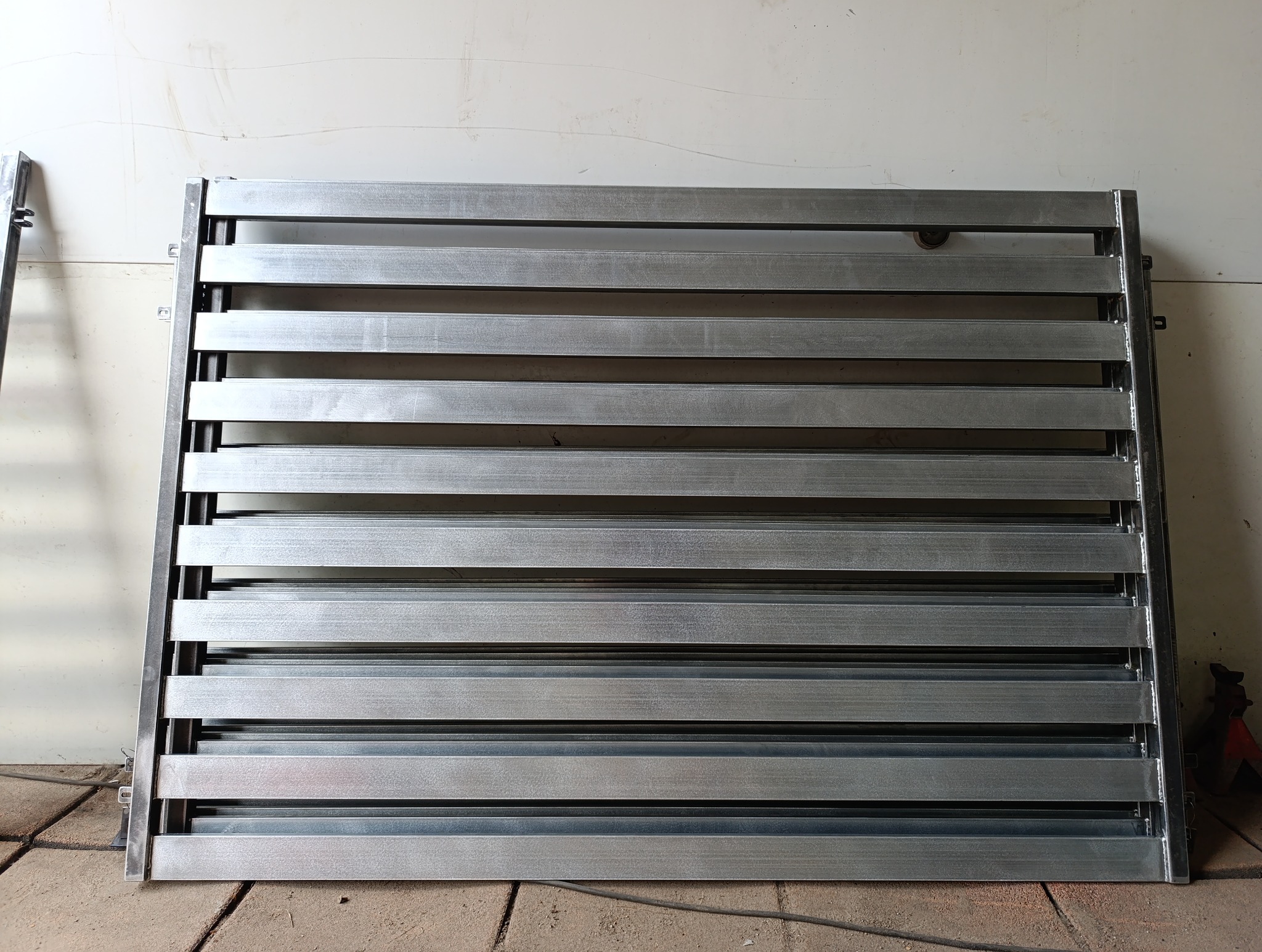 Metalowy segment ogrodzeniowy z poziomymi lamelami, surowy, przed montażem. Widoczne detale konstrukcyjne i elementy mocujące. Szary odcień metalu.