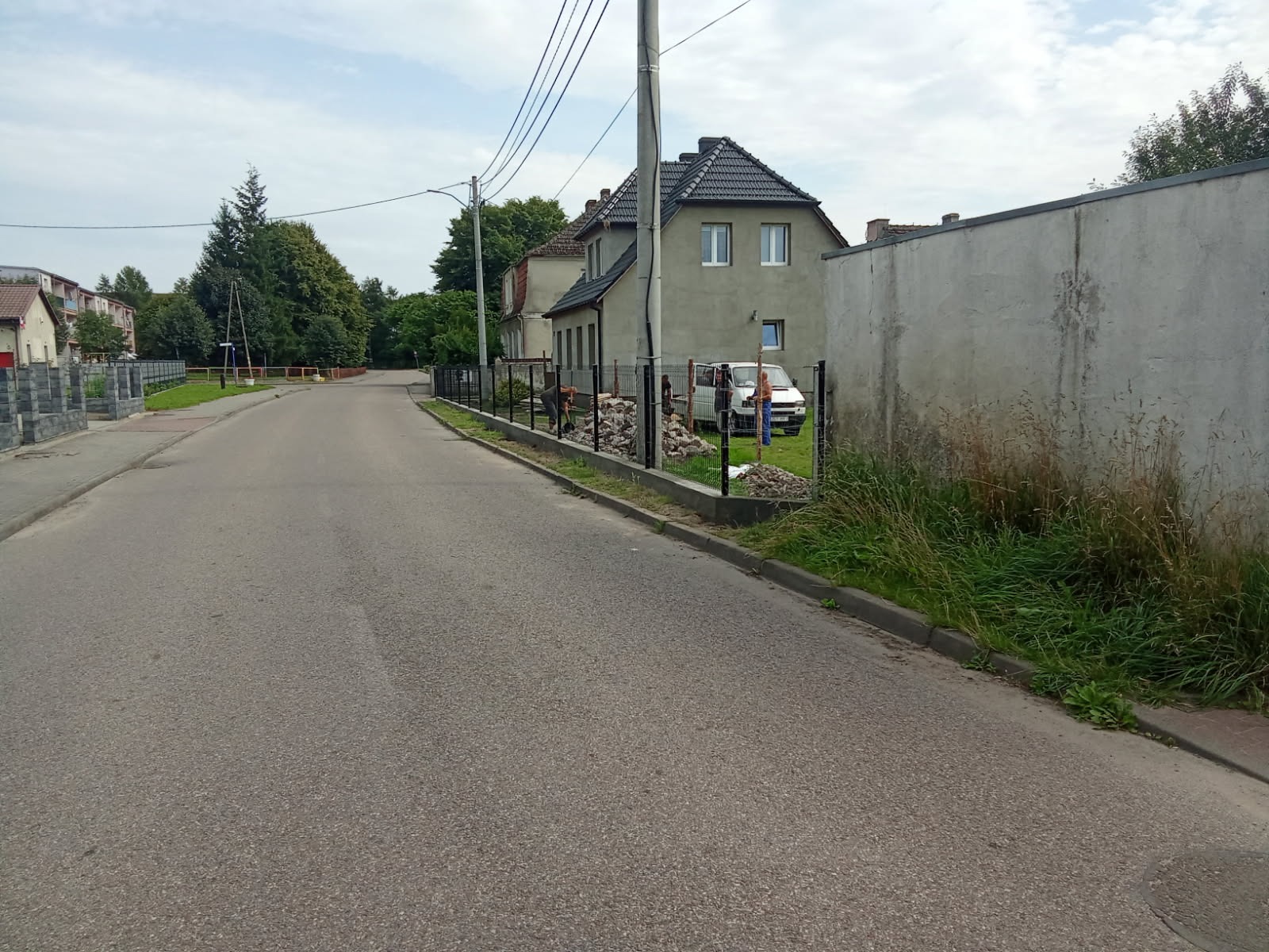 Montaż ogrodzenia panelowego przy drodze; widoczne słupki i panele, w tle dom i zaparkowany samochód. Prace w toku, przygotowanie podłoża.