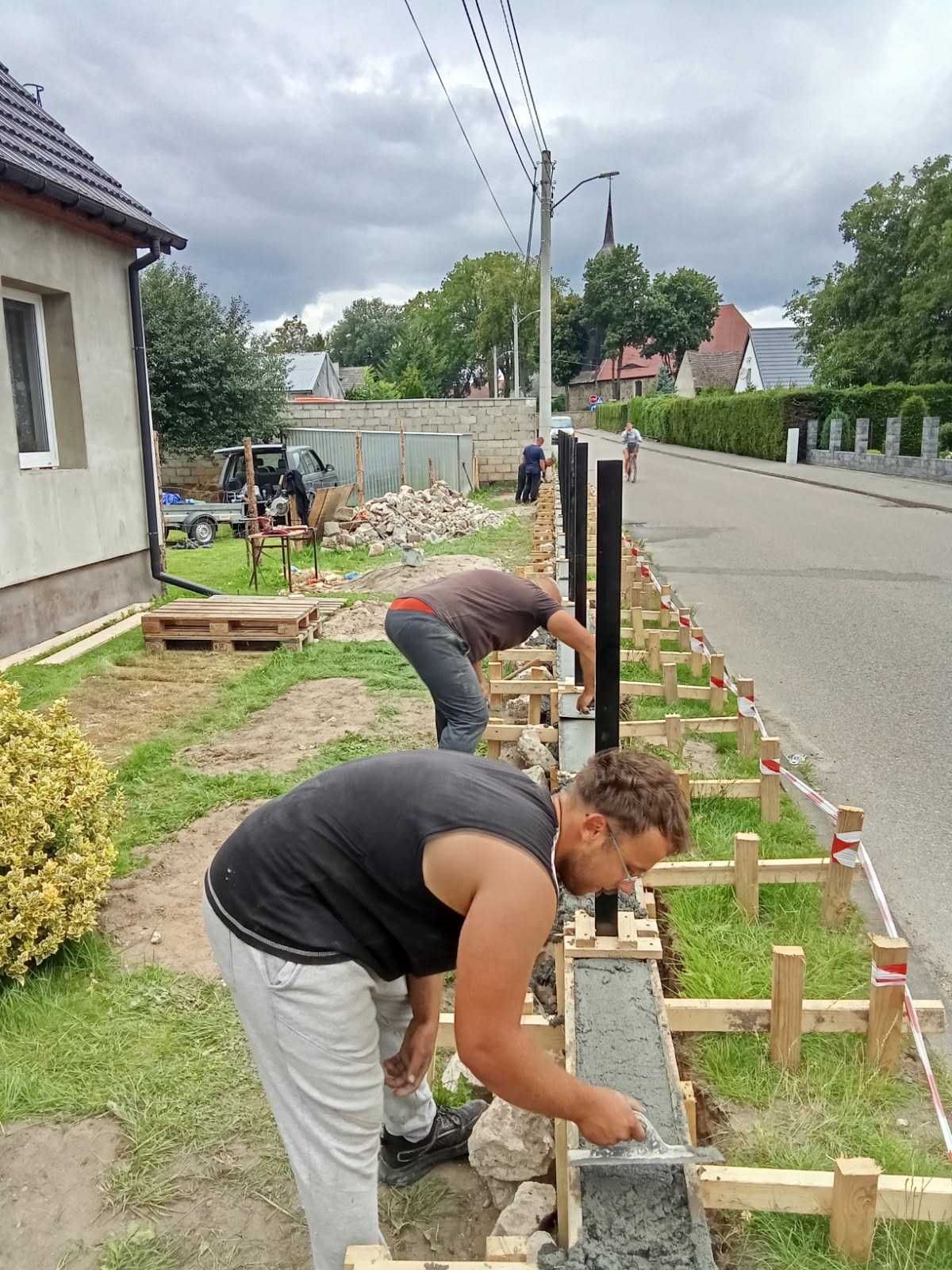 Dwóch mężczyzn w trakcie betonowania fundamentu pod ogrodzenie z czarnymi słupkami. Widoczne drewniane szalunki i narzędzia. Praca w toku, obok droga i dom.