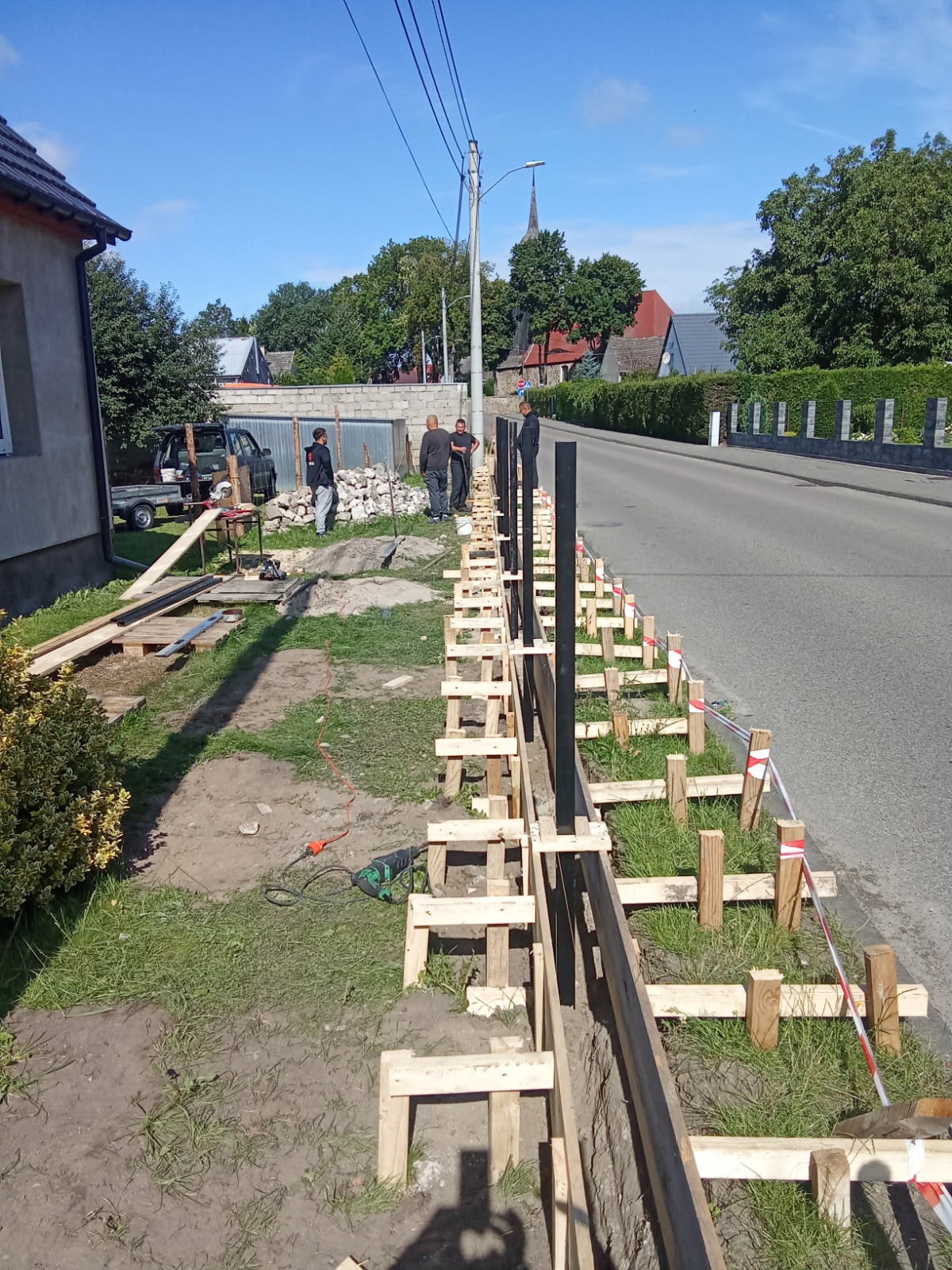 Wykop pod fundament ogrodzenia z drewnianym szalunkiem i czarnymi słupkami, widoczni robotnicy, narzędzia i materiały budowlane. Praca w toku, widok na ulicę i zabudowania.