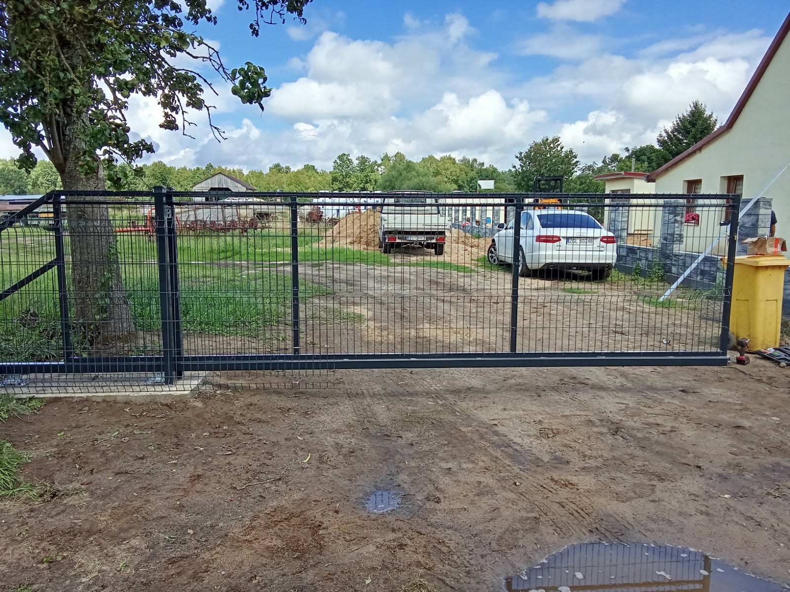 Czarna, przesuwna brama wjazdowa z siatki panelowej na posesji z budynkiem i samochodami w tle, widok z zewnątrz. Słoneczny dzień z widocznym niebem.
