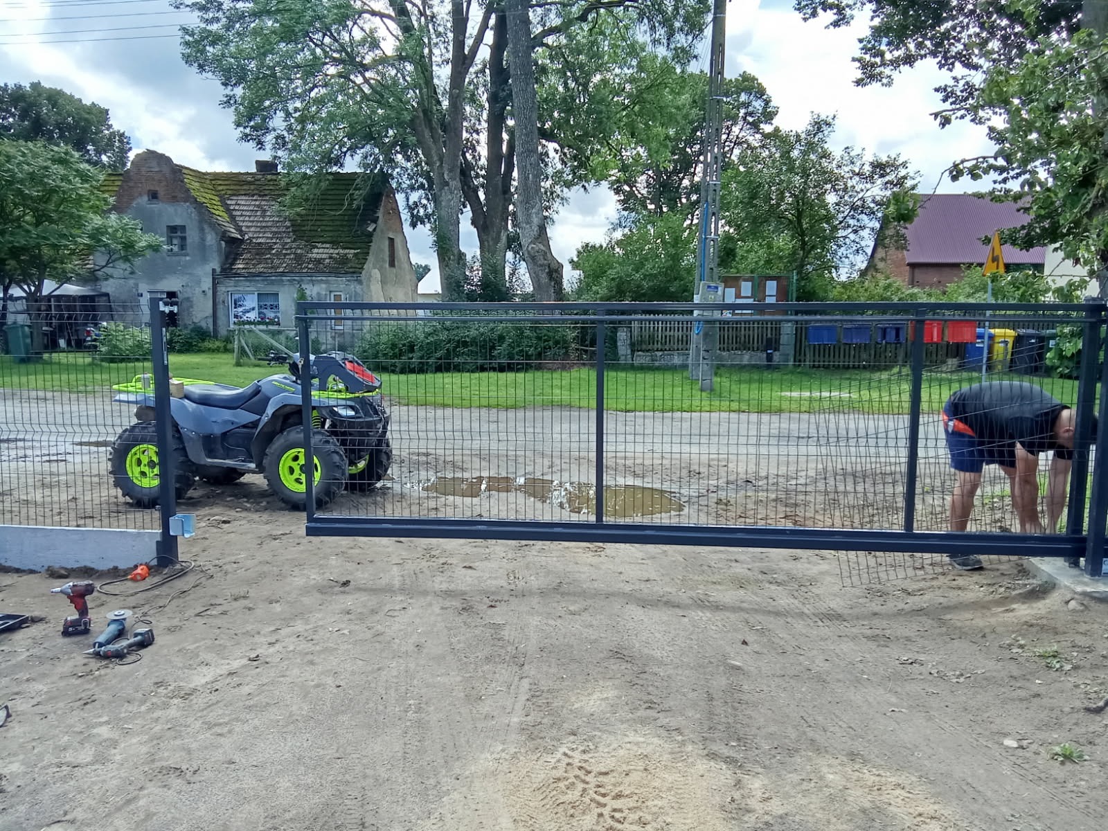 Montaż przesuwnej bramy wjazdowej z siatki na posesji. Obok quad i narzędzia. Prace instalacyjne w toku, widoczna sylwetka montera. Tło: domy i zieleń.