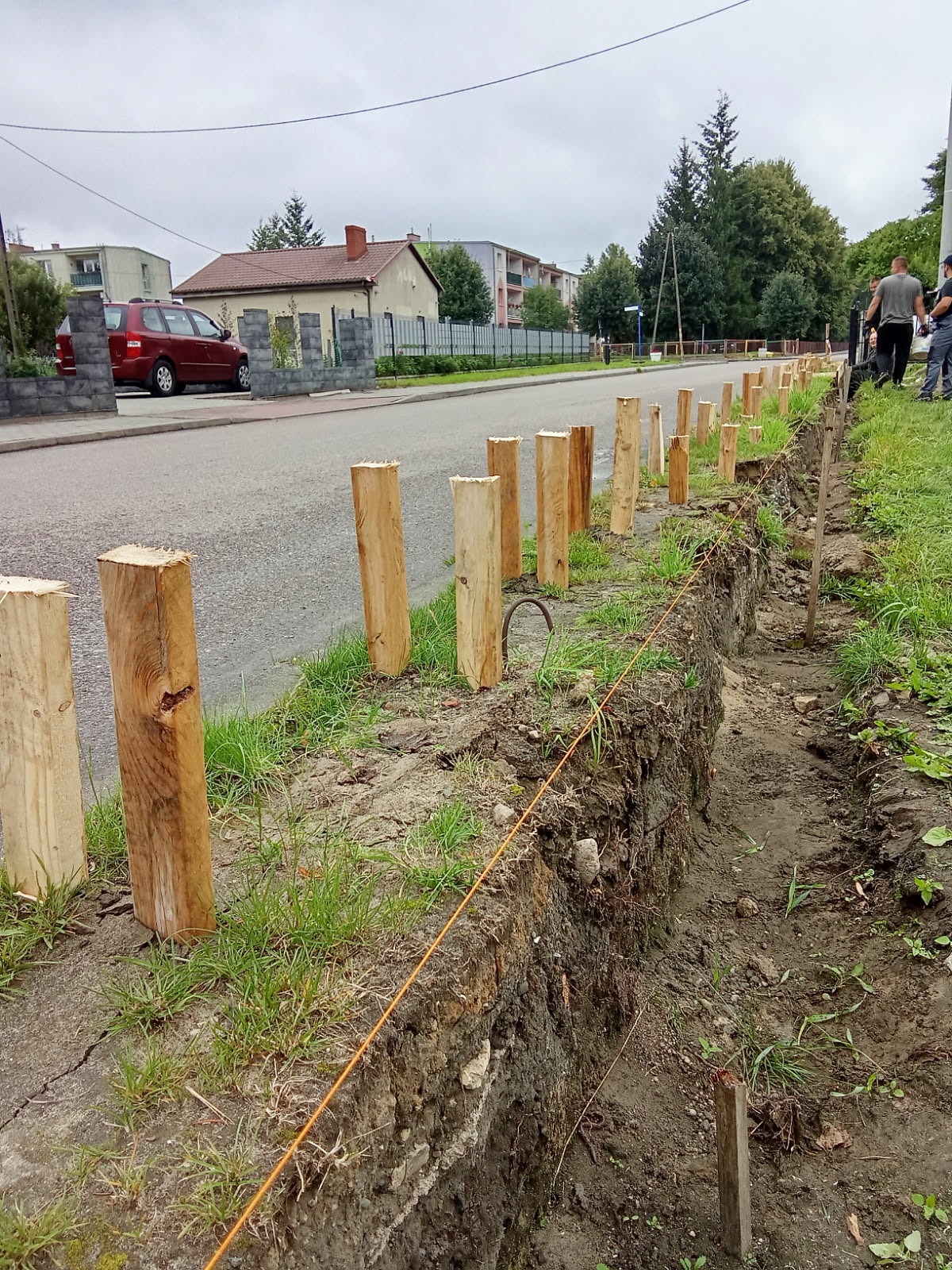 Etap przygotowania podłoża pod montaż ogrodzenia: wykopany rów, wbite drewniane słupki wyrównane sznurkiem. W tle zabudowa mieszkalna i fragmenty ulicy.