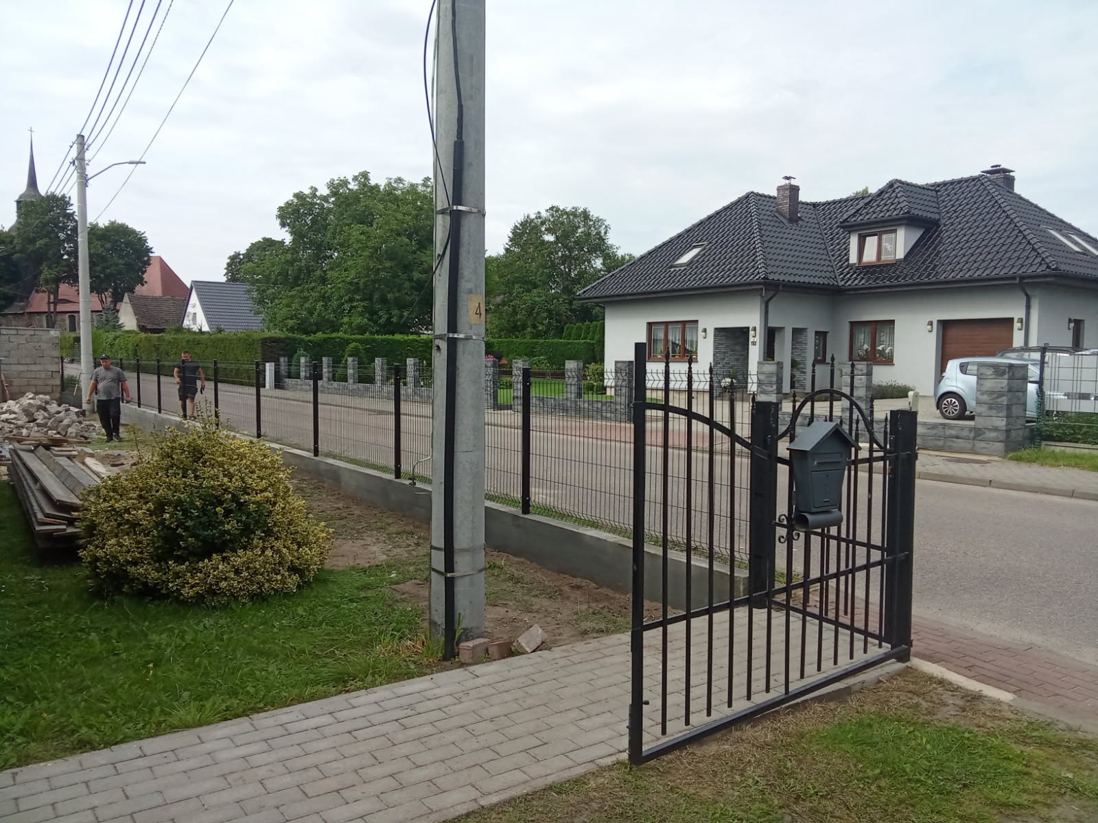 Ukończony montaż ogrodzenia panelowego z bramką i skrzynką na listy. W tle widoczny dom jednorodzinny i dwóch mężczyzn przy pracy.