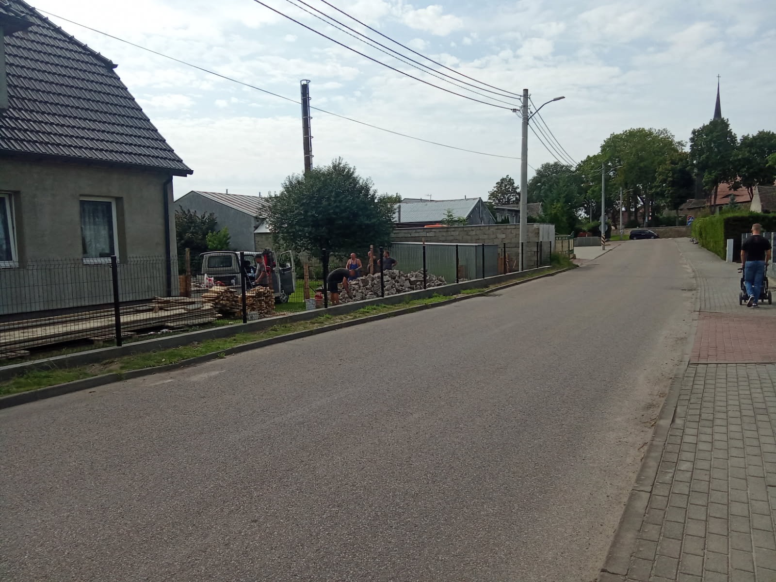 Montaż ogrodzenia panelowego na posesji w trakcie realizacji. Widoczne panele, słupki i ekipa montażowa przy pracy. Elementy ogrodzenia składowane obok.