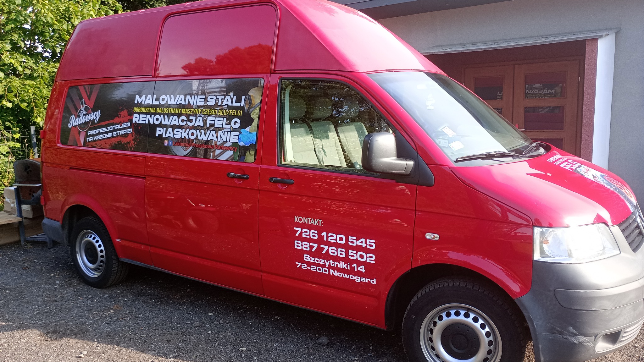 Czerwony bus z reklamą firmy oferującej malowanie stali, renowację felg i piaskowanie, zaparkowany na żwirowym podłożu przed budynkiem.