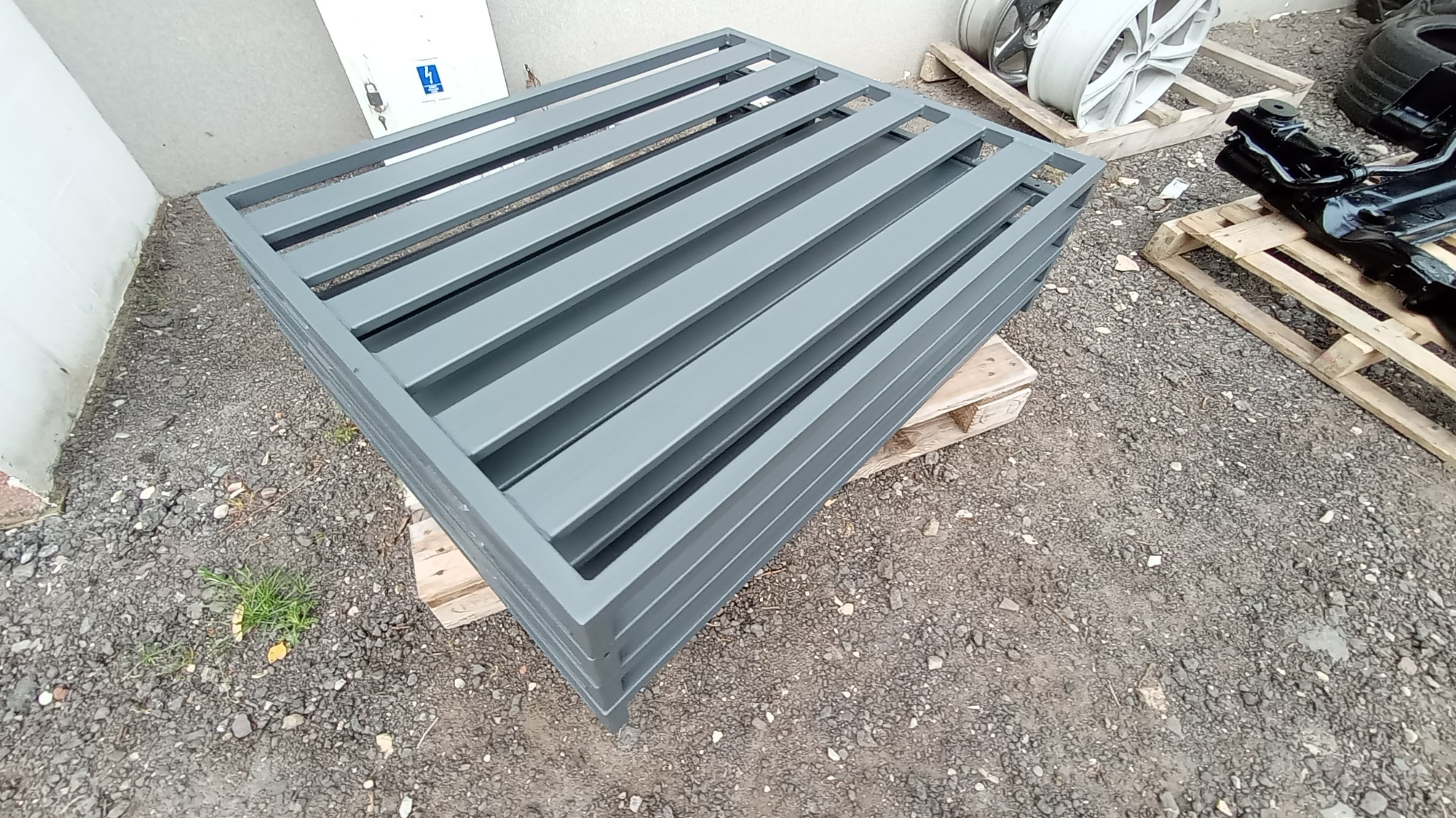 Stos pomalowanych na szaro metalowych elementów kratowych, ułożonych na palecie na żwirowym podłożu, z fragmentami felg i zawieszenia samochodowego w tle.