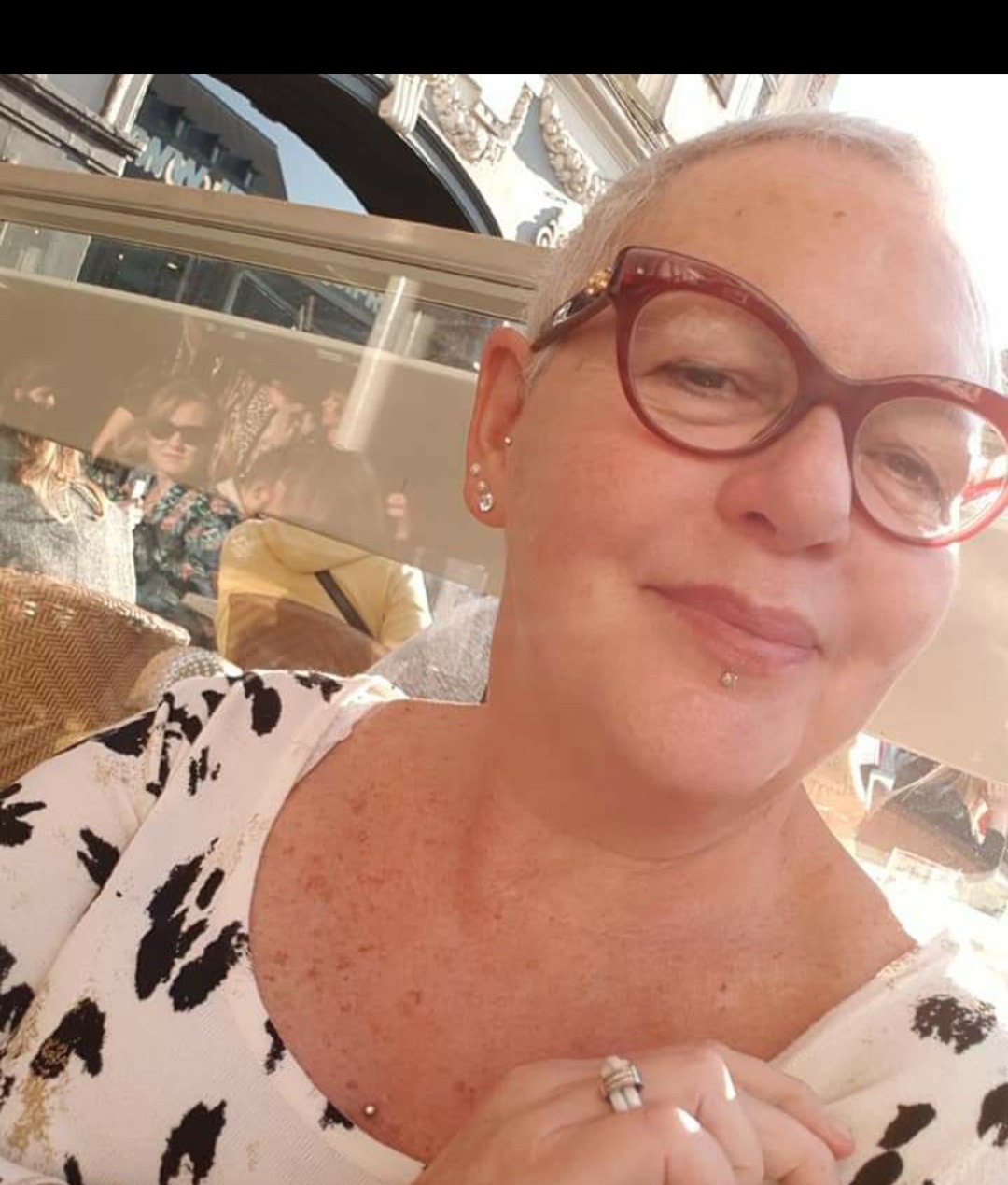 Portret kobiety w okularach w kratkę, z piercingiem na wardze i kolczykami, siedzącej w kawiarni z odbiciem w szybie