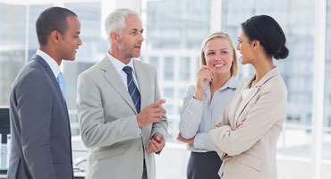 Grupa czterech uśmiechniętych, elegancko ubranych osób (dwóch mężczyzn i dwie kobiety) rozmawia w jasnym, nowoczesnym biurze z dużymi oknami.