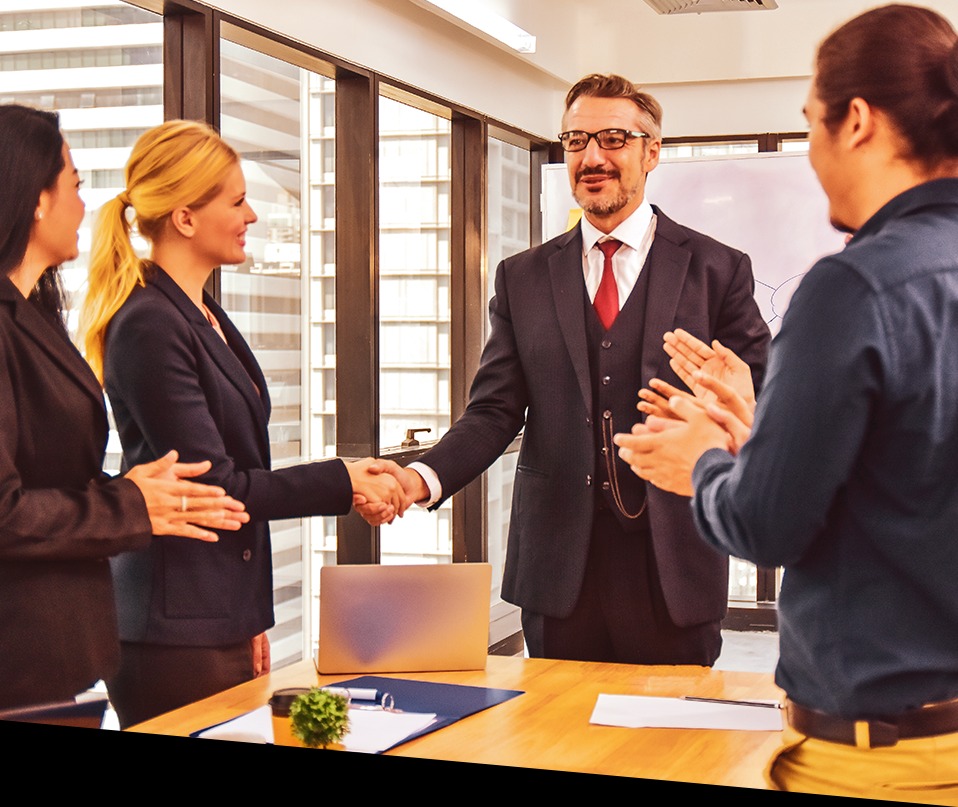 Biznesmen w garniturze ściska dłoń kobiecie podczas spotkania biznesowego, z laptopem i dokumentami na stole, a inni współpracownicy klaszczą w tle.