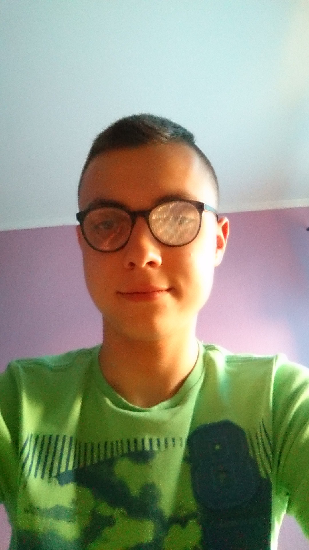 Portret młodego mężczyzny w okularach z zieloną koszulką na tle fioletowej ściany i białego sufitu.