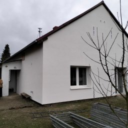 DMBAU SPÓŁKA Z OGRANICZONĄ ODPOWIEDZIALNOŚCIĄ - Biały dom po renowacji elewacji, z widoczną osobą i elementami otoczenia, takimi jak drzewo bez liści i drabiny. Świeżo otynkowana fasada budynku.