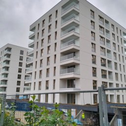 DMBAU SPÓŁKA Z OGRANICZONĄ ODPOWIEDZIALNOŚCIĄ - Nowoczesny budynek wielorodzinny z balkonami i dużymi oknami, widziany z dołu, z elementami placu budowy w tle i siatką ogrodzeniową na pierwszym planie.