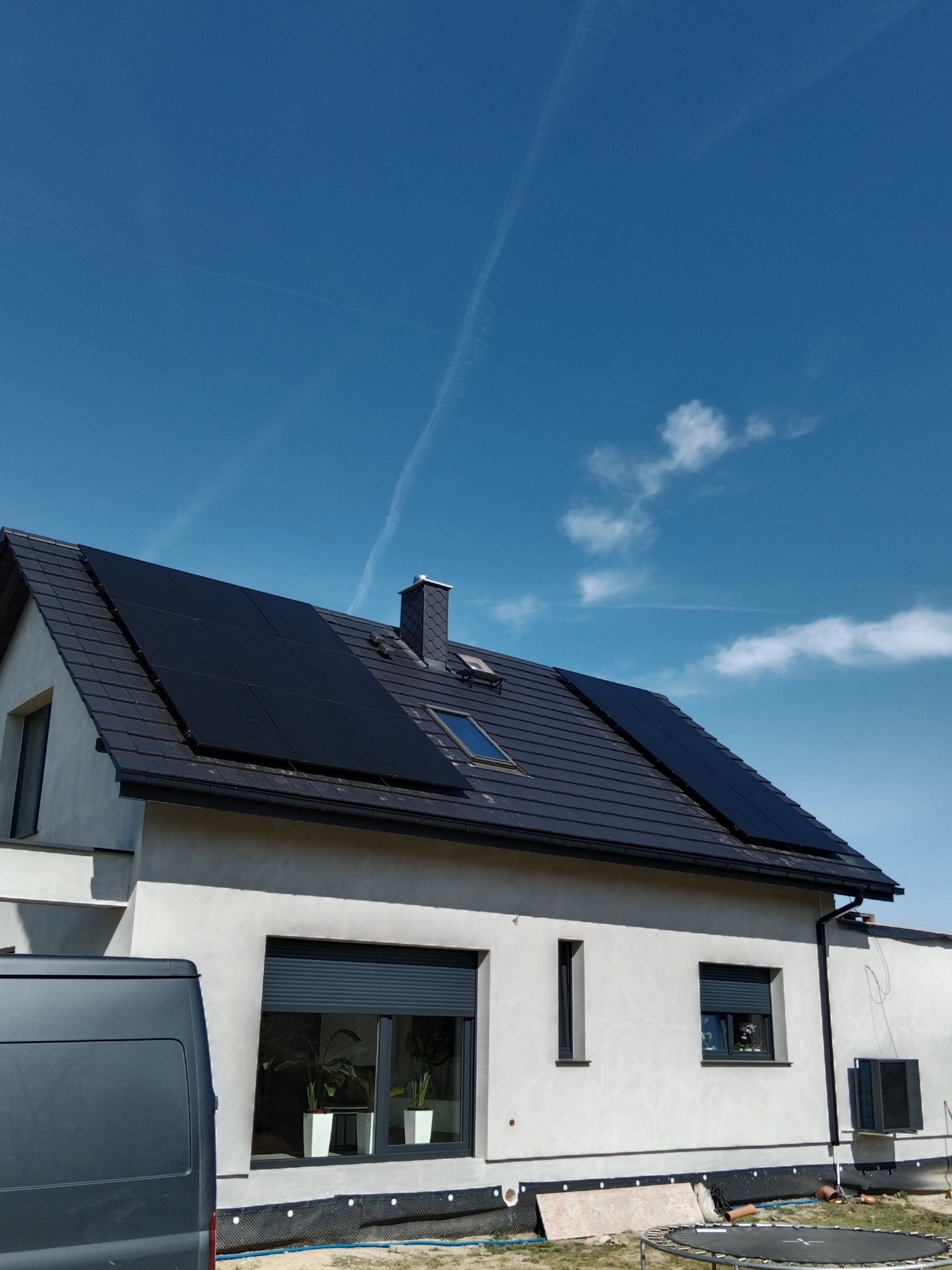 Nowoczesny dom z szarym dachem i panelami słonecznymi na dachu, widok z dołu w słoneczny dzień z lekkimi chmurami i śladami kondensacji po samolotach.