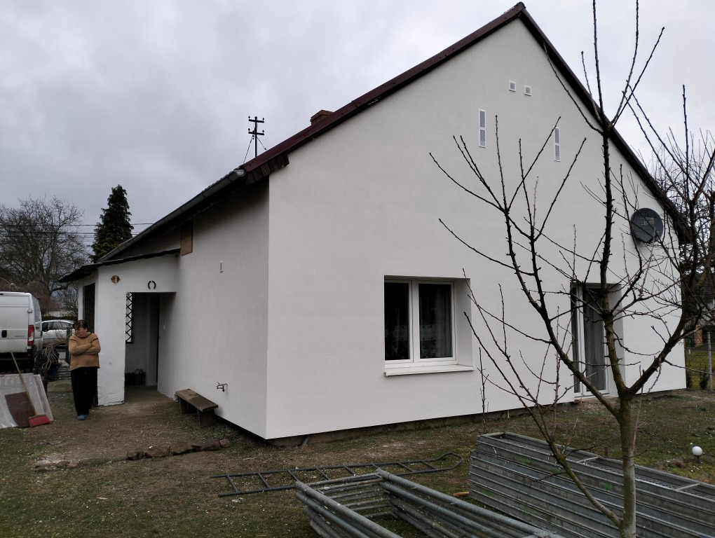 Biały dom po renowacji elewacji, z widoczną osobą i elementami otoczenia, takimi jak drzewo bez liści i drabiny. Świeżo otynkowana fasada budynku.