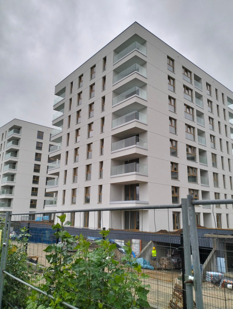 Nowoczesny budynek wielorodzinny z balkonami i dużymi oknami, widziany z dołu, z elementami placu budowy w tle i siatką ogrodzeniową na pierwszym planie.