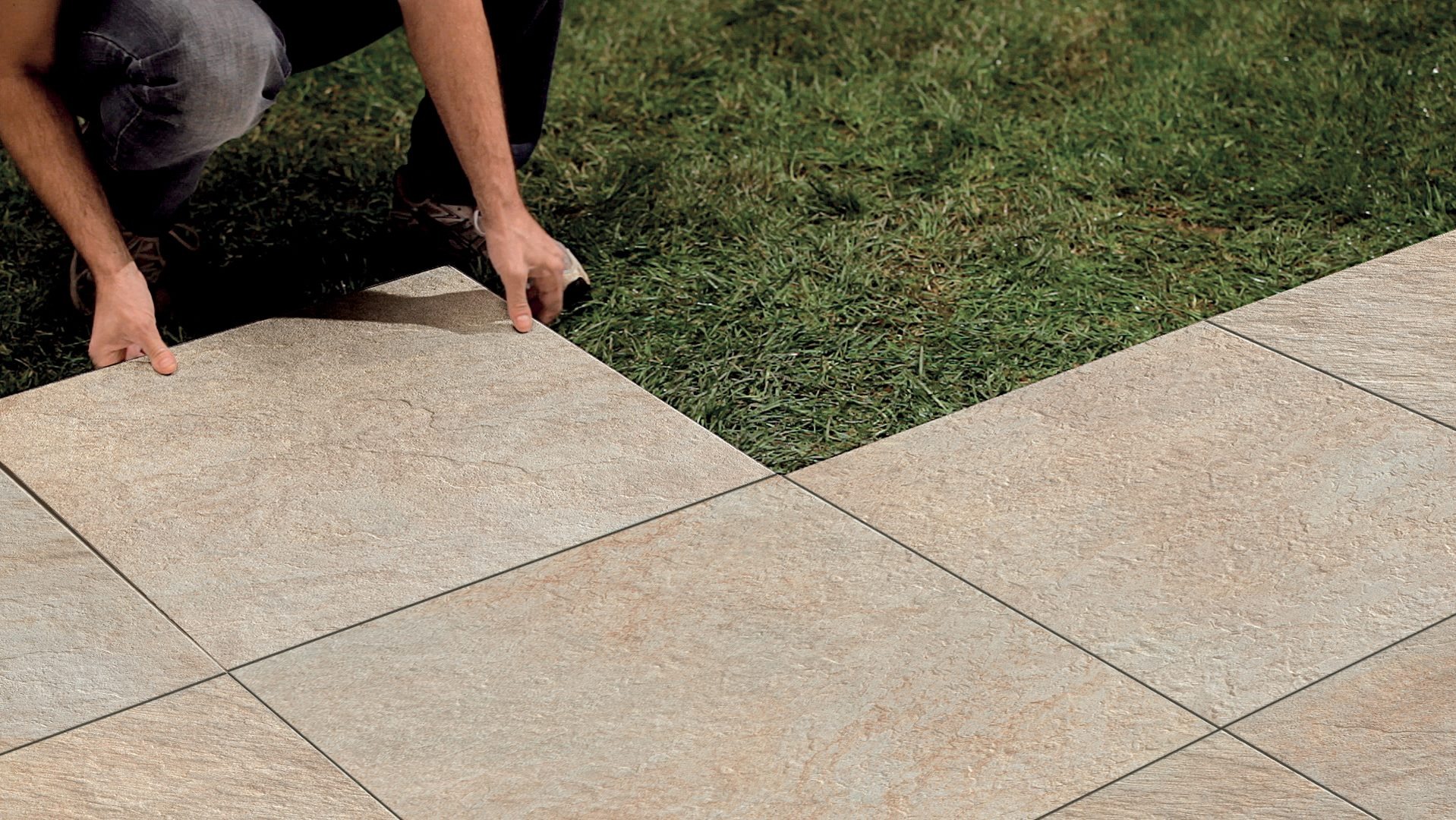 Układanie beżowych płyt chodnikowych na trawie, widoczne ręce układające ostatni element, tworząc geometryczny wzór na styku z zielenią.