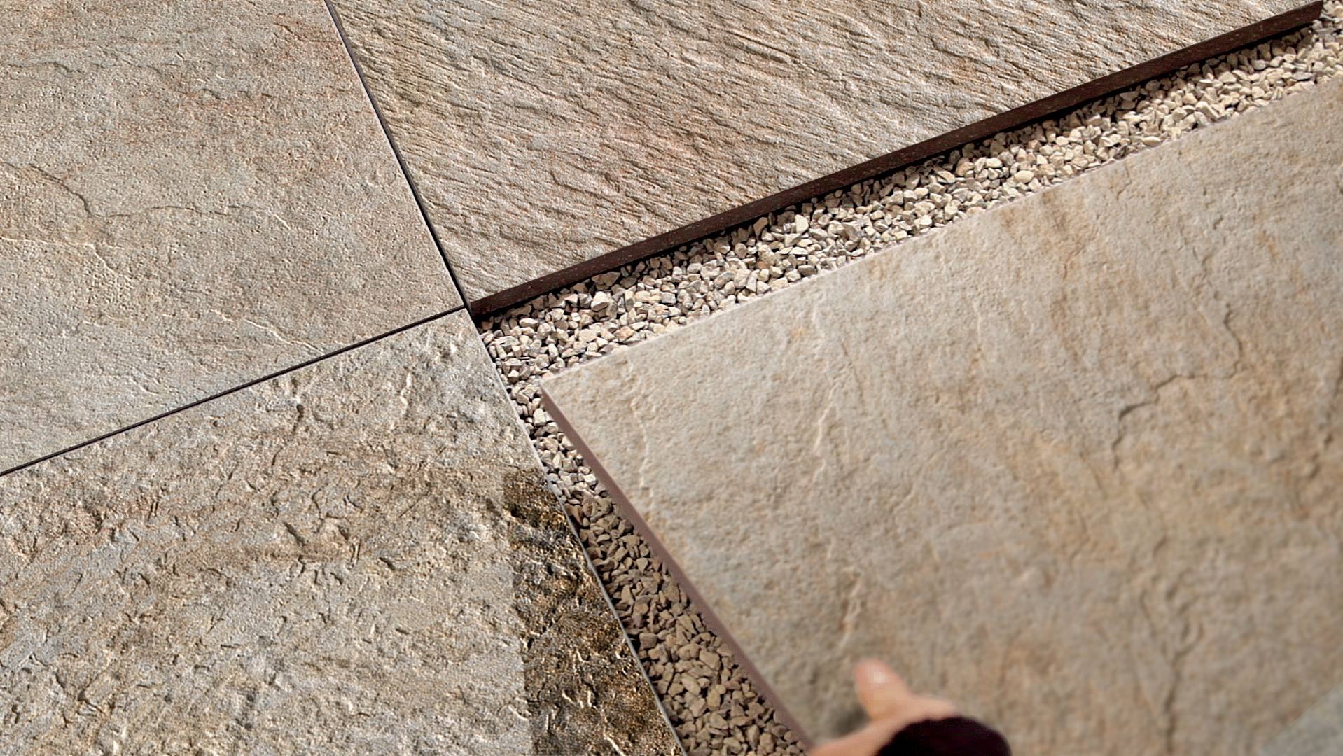 Detale ułożenia beżowych płytek podłogowych imitujących kamień, z brązową listwą i drobnymi kamyczkami dekoracyjnymi w fugach.