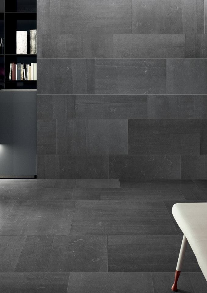 Minimalistyczne wnętrze z ciemnoszarą, wielkoformatową płytką gresową na podłodze i ścianie, uzupełnione czarnym regałem z książkami i fragmentem białego krzesła.