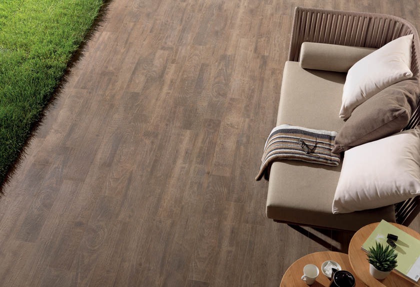 Brązowa podłoga tarasowa imitująca drewno, z widoczną fakturą słojów, sąsiadująca z trawnikiem. Na podłodze leżanka z beżowymi poduszkami i stoliki kawowe z akcesoriami.