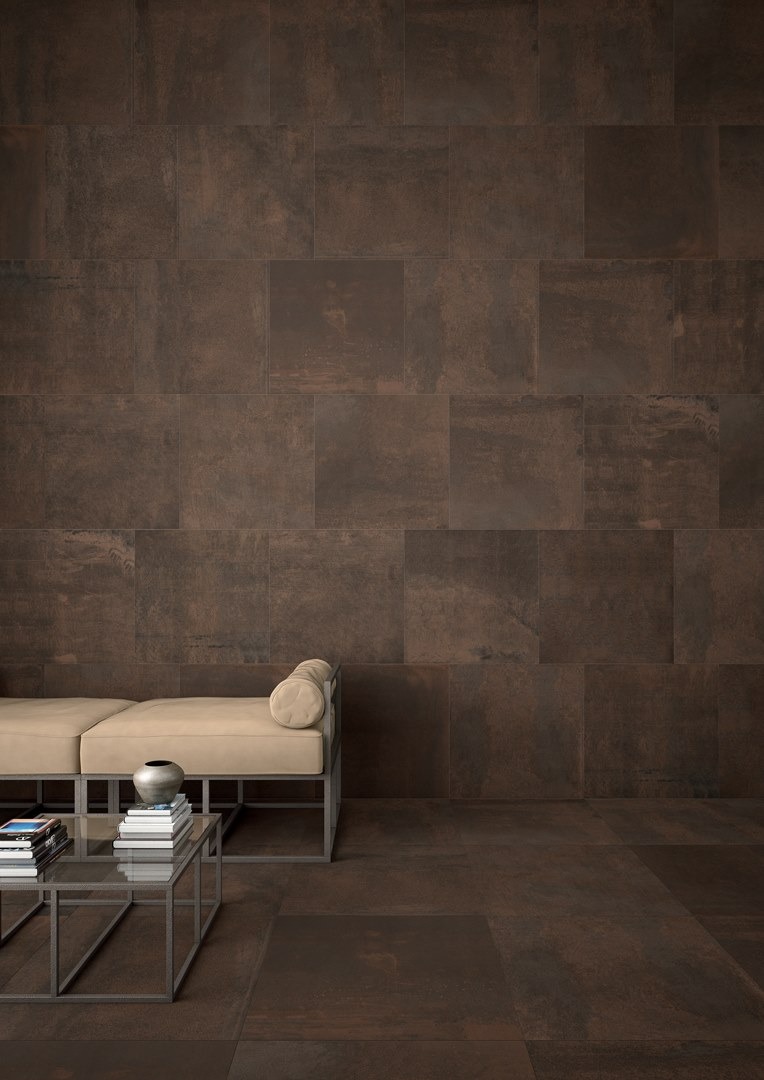 Minimalistyczny salon z ścianą i podłogą wyłożoną dużymi, brązowymi płytkami imitującymi metal, jasna sofa z metalową ramą i szklany stolik kawowy z książkami i wazonem.