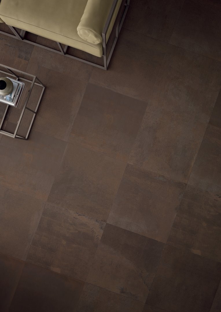 Brązowe, matowe płytki podłogowe ułożone w kwadraty, widoczne z góry, z fragmentem sofy i stolika kawowego w tle.