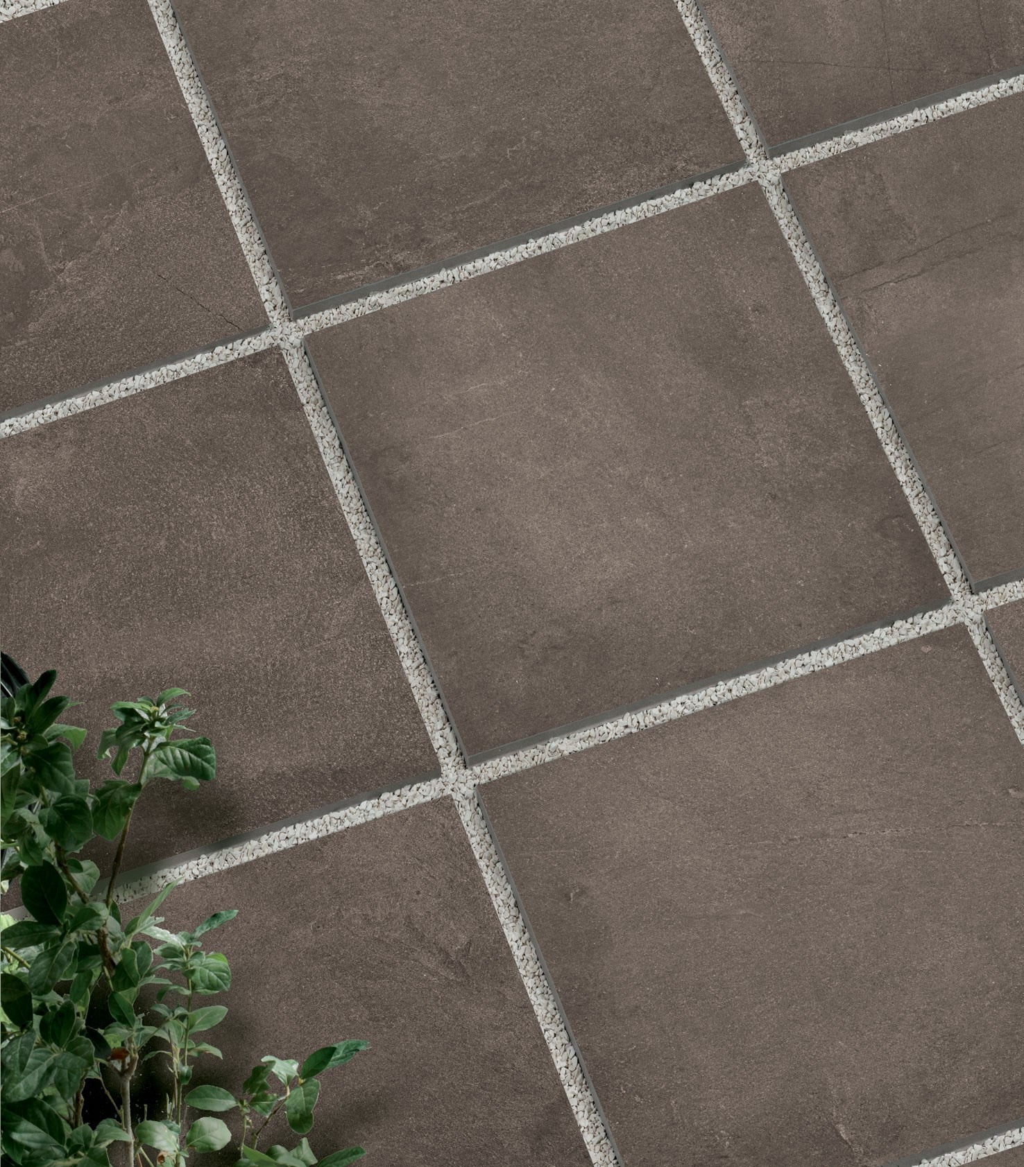 Brunatne płytki podłogowe ułożone w kwadraty z jasnymi kamyczkami w fugach, widok z góry z fragmentem zielonej rośliny w lewym dolnym rogu.