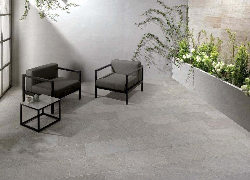 Jasnoszare płytki podłogowe imitujące kamień na tarasie z dwoma fotelami o metalowych ramach, stolikiem kawowym i roślinami pnącymi się po ścianie.