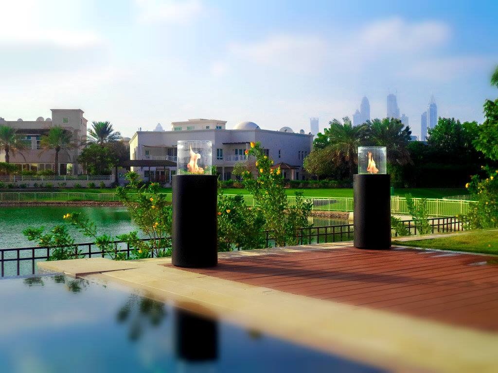 Dwa zewnętrzne kominki gazowe na tarasie z drewnianą podłogą, widok na jezioro, budynki mieszkalne i odległe wieżowce w Dubaju.