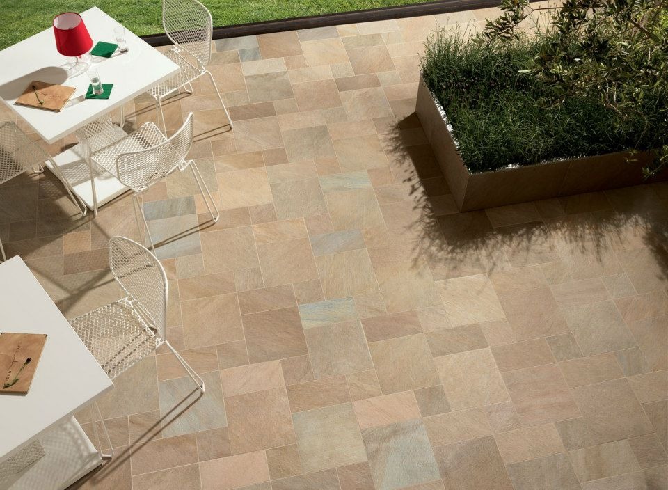 Beżowa, prostokątna posadzka z płytek na tarasie, uzupełniona białymi krzesłami z metalowej siatki, białymi stolikami i donicą z zielenią.