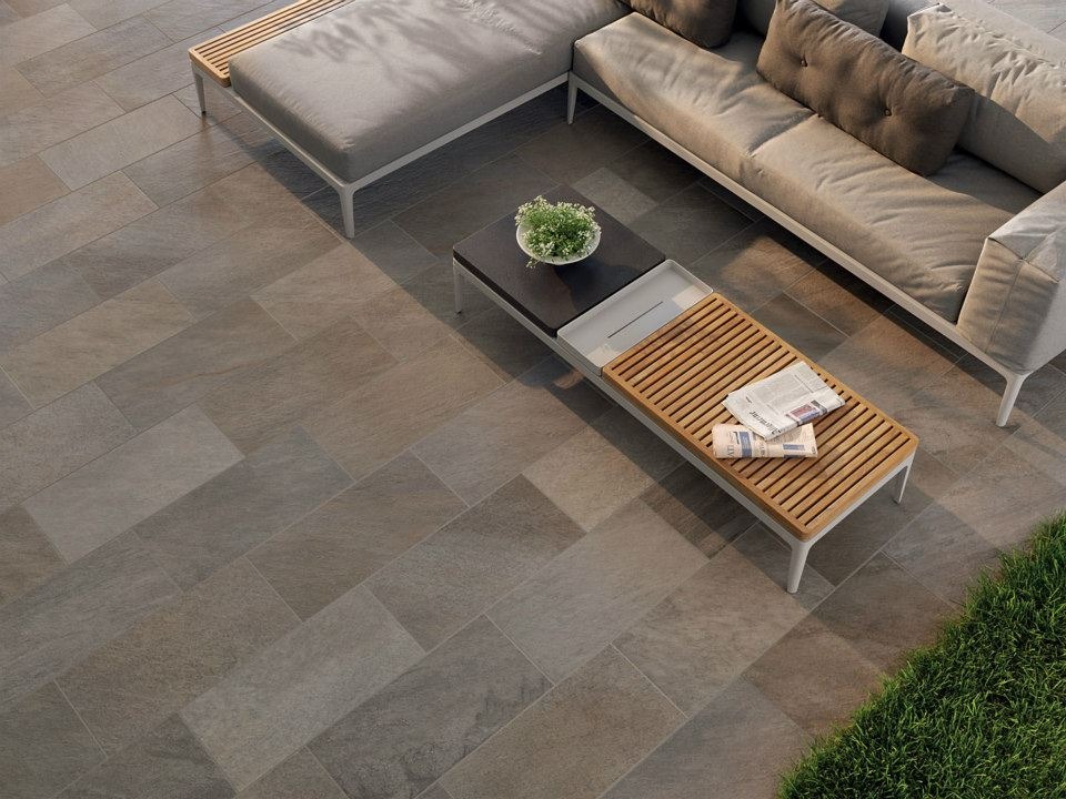 Zewnętrzny taras wyłożony płytkami imitującymi kamień, z nowoczesnymi meblami ogrodowymi: sofa z poduszkami, stolik kawowy z drewnianym blatem i doniczką z kwiatami, gazety leżące na blacie