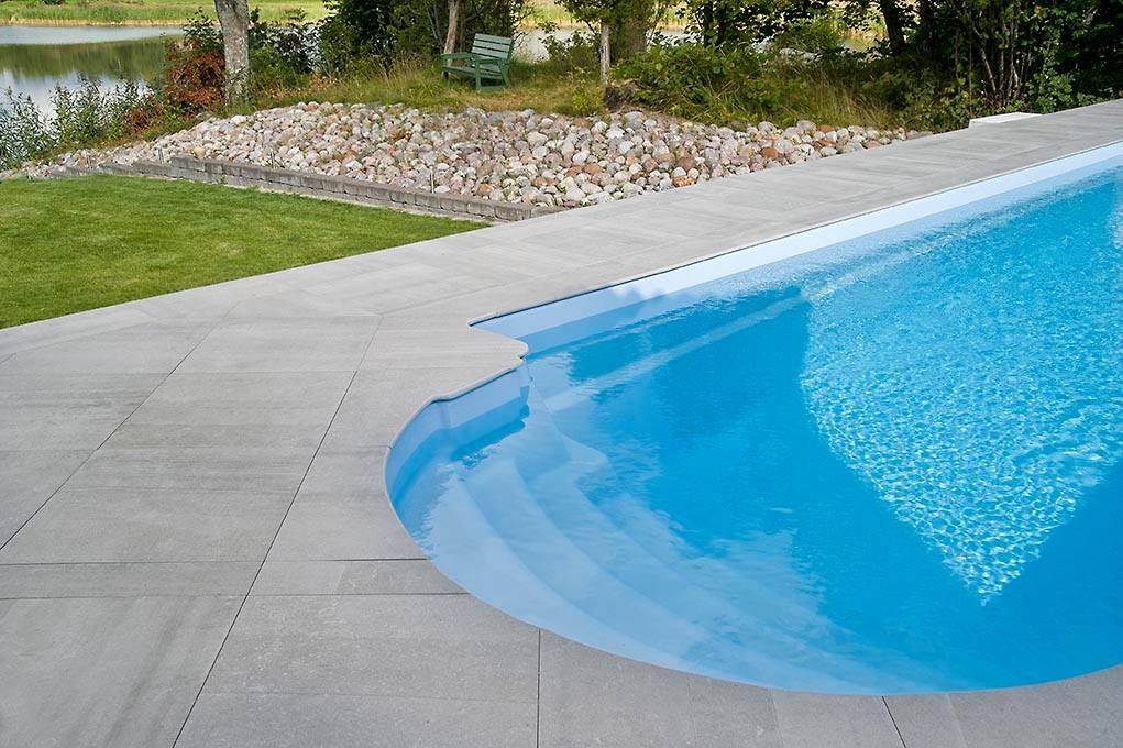 Nowoczesny basen z szarymi płytami tarasowymi, widok na jezioro i zielony brzeg z kamieniami.