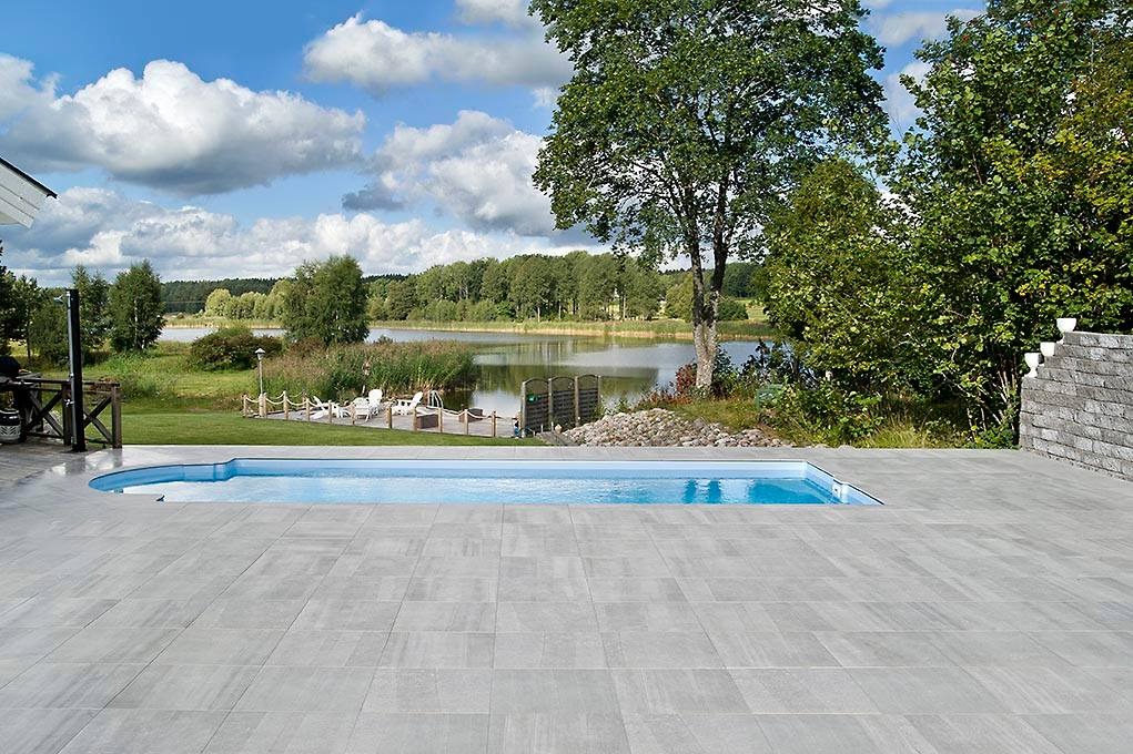 Nowoczesny basen z szarymi płytami tarasowymi, widok na jezioro i las w tle, błękitne niebo z białymi chmurami.